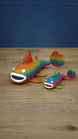 Two rainbow Palia Prismfish 3D printed figures displayed on a wooden tabletop, featuring bright rainbow colouring and white eyes.