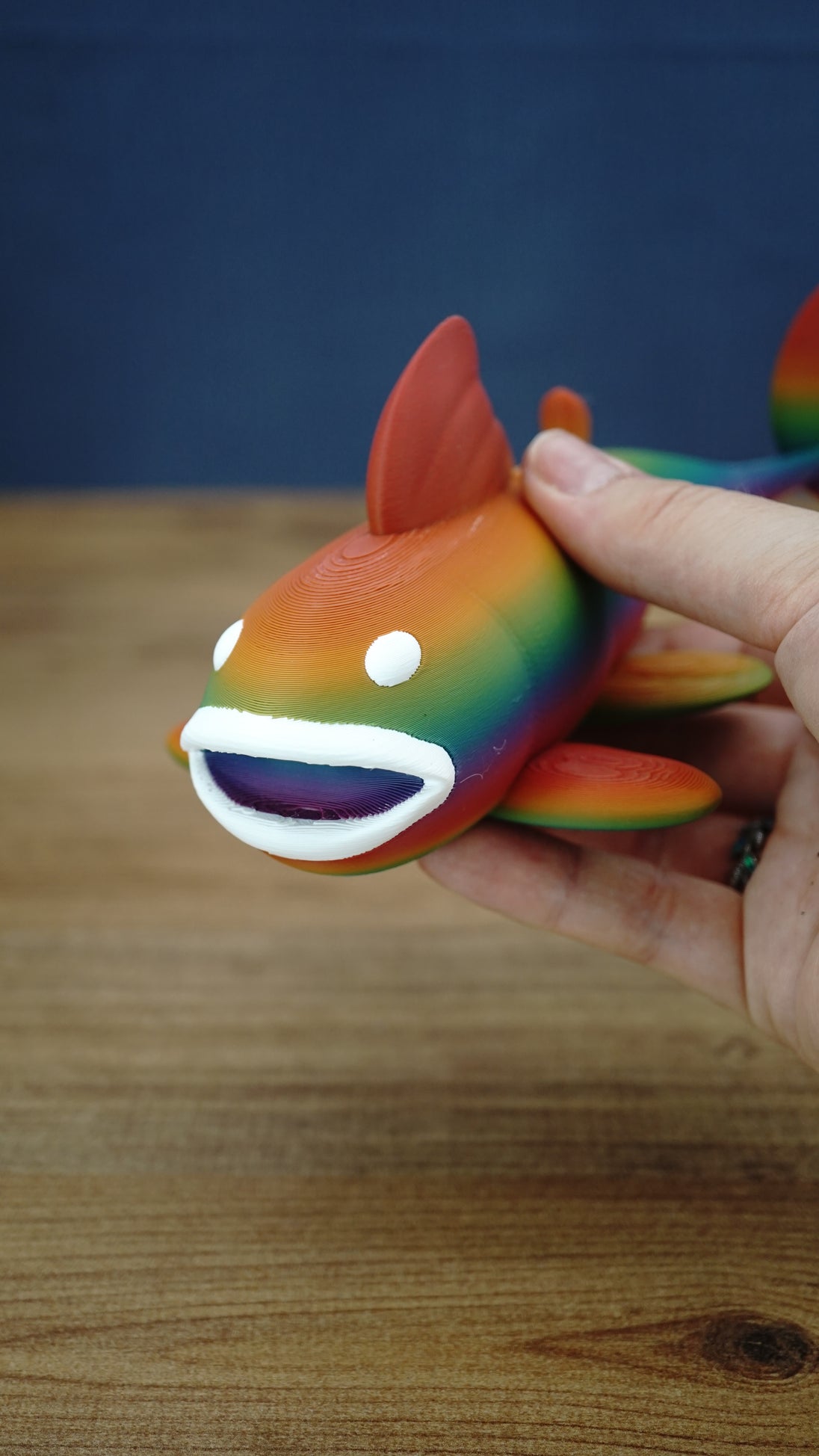 Close-up of a rainbow Palia Prismfish 3D printed figure being held, showing its open white mouth, white eyes, and colourful body.