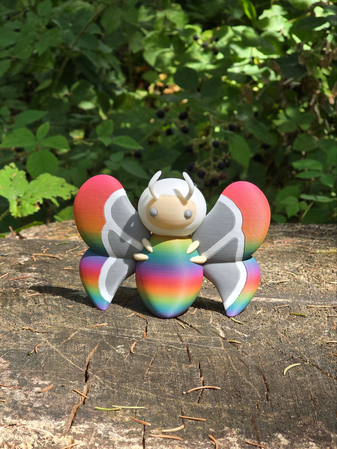3D printed Palia Rainbow-Tipped Butterfly plush figure with rainbow body and red-tipped wings sitting on a tree stump in the forest.