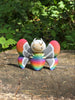 3D printed Palia Rainbow-Tipped Butterfly plush figure with rainbow body and red-tipped wings sitting on a tree stump in the forest.