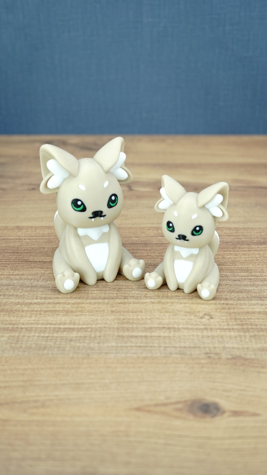 Two 3D printed Palia-inspired Sandi the Palcat plush figures in sandy beige with white accents, green eyes, and white paw pads on a wooden tabletop.