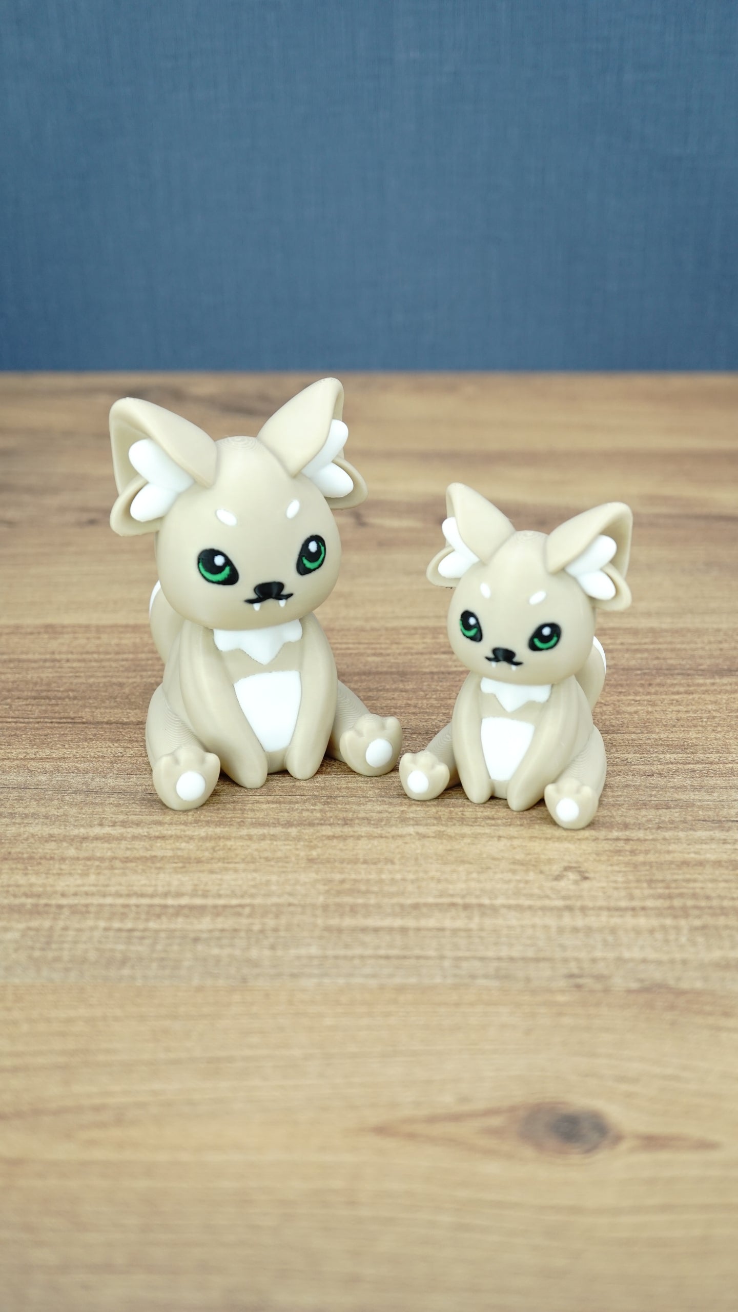 Two 3D printed Palia-inspired Sandi the Palcat plush figures in sandy beige with white accents, green eyes, and white paw pads on a wooden tabletop.