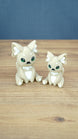 Two 3D printed Palia-inspired Sandi the Palcat plush figures in sandy beige with white accents, green eyes, and white paw pads on a wooden tabletop.