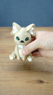 Handheld close-up of a 3D printed Sandi the Palcat plush figure with sandy beige body, white accents, green eyes, tiny fangs, and white paw pads.