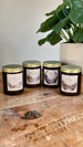 Four Palia scented soy wax candles—Royal Highlands, Bahari Bay, Elderwood and Kilima Valley—in amber jars with gold lids on a wooden table.