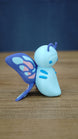 Side view of the Palia Shimmerfly Plush inspired collectible showing its rounded blue body, curled antennae and layered purple butterfly wings