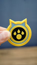 Palworld Dog Coin replica held between fingers to demonstrate its compact 4cm size and detailed yellow, light blue and black design.