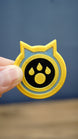 Palworld Dog Coin replica held between fingers to demonstrate its compact 4cm size and detailed yellow, light blue and black design.