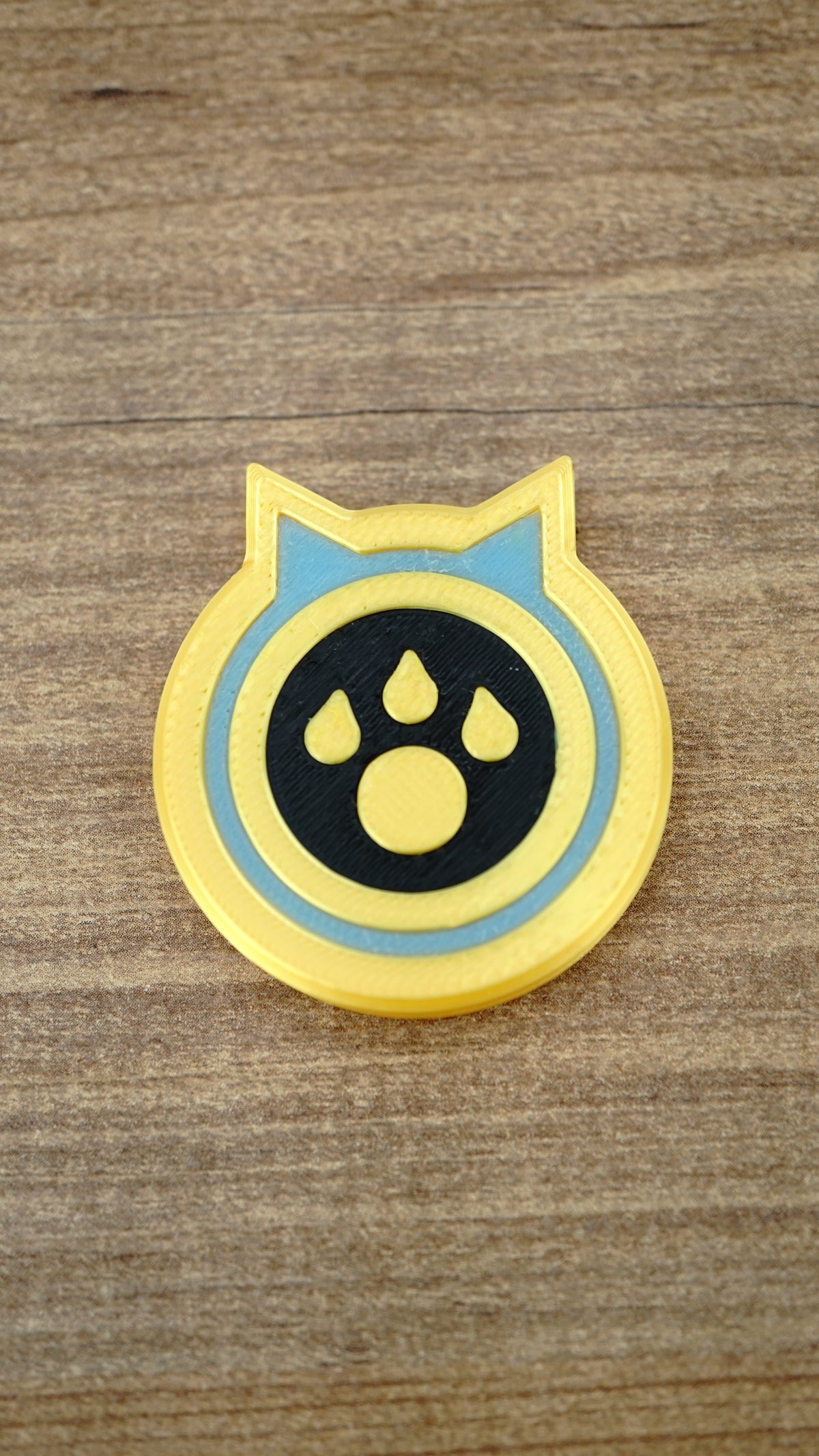 Single Palworld Dog Coin replica displayed face-up on wood, featuring a yellow outer edge, light blue ring and black paw emblem centre.