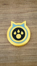 Single Palworld Dog Coin replica displayed face-up on wood, featuring a yellow outer edge, light blue ring and black paw emblem centre.