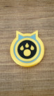 Single Palworld Dog Coin replica displayed face-up on wood, featuring a yellow outer edge, light blue ring and black paw emblem centre.