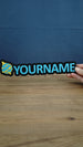Blue and yellow personalised Palworld Sphere nameplate held in hand to show its 25cm scale, with raised YOURNAME lettering and a black display base.