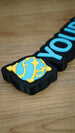 Close-up of the blue and yellow Sphere emblem on a personalised Palworld nameplate, showing the layered detail and black base on a wooden surface.