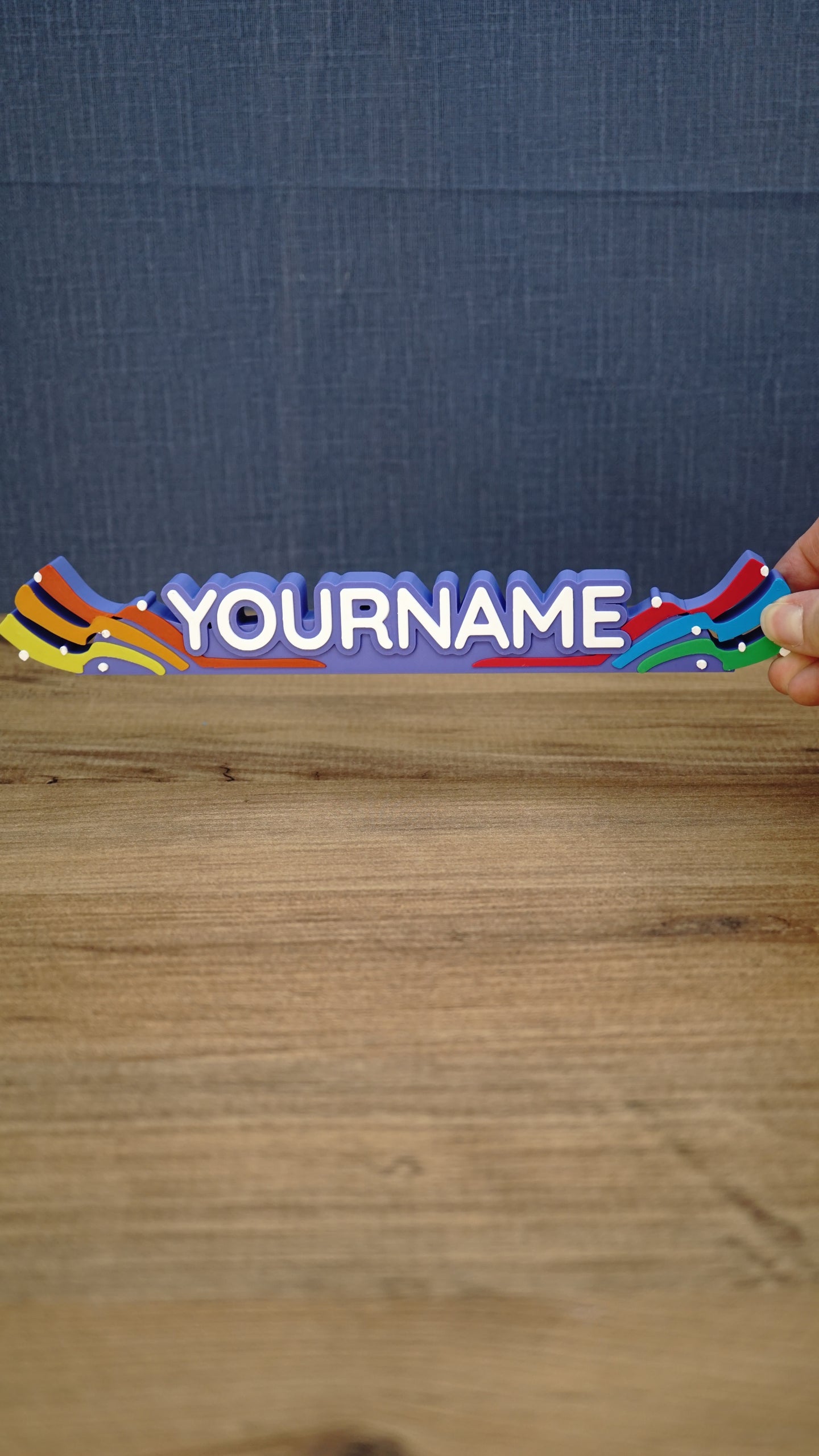 Front view of a 25 cm personalized Palia Prismatic Pride nameplate held above a wooden table, with rainbow ends and bold white lettering.