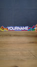 Front view of a 25 cm personalized Palia Prismatic Pride nameplate held above a wooden table, with rainbow ends and bold white lettering.
