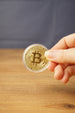 Hand holding a gold physical Bitcoin replica in a clear protective case, showing the raised Bitcoin logo and engraved coin details.