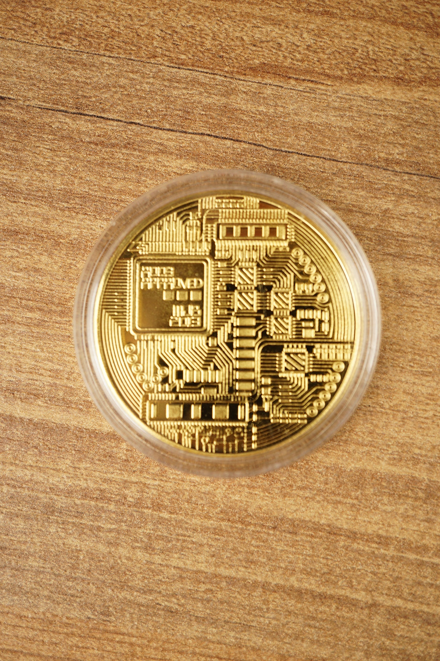 Gold physical Bitcoin replica in a clear protective capsule, shown from above on a wooden tabletop with detailed circuit-style engraving.
