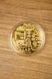 Gold physical Bitcoin replica in a clear protective capsule, shown from above on a wooden tabletop with detailed circuit-style engraving.