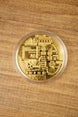 Gold physical Bitcoin replica in a clear protective capsule, shown from above on a wooden tabletop with detailed circuit-style engraving.