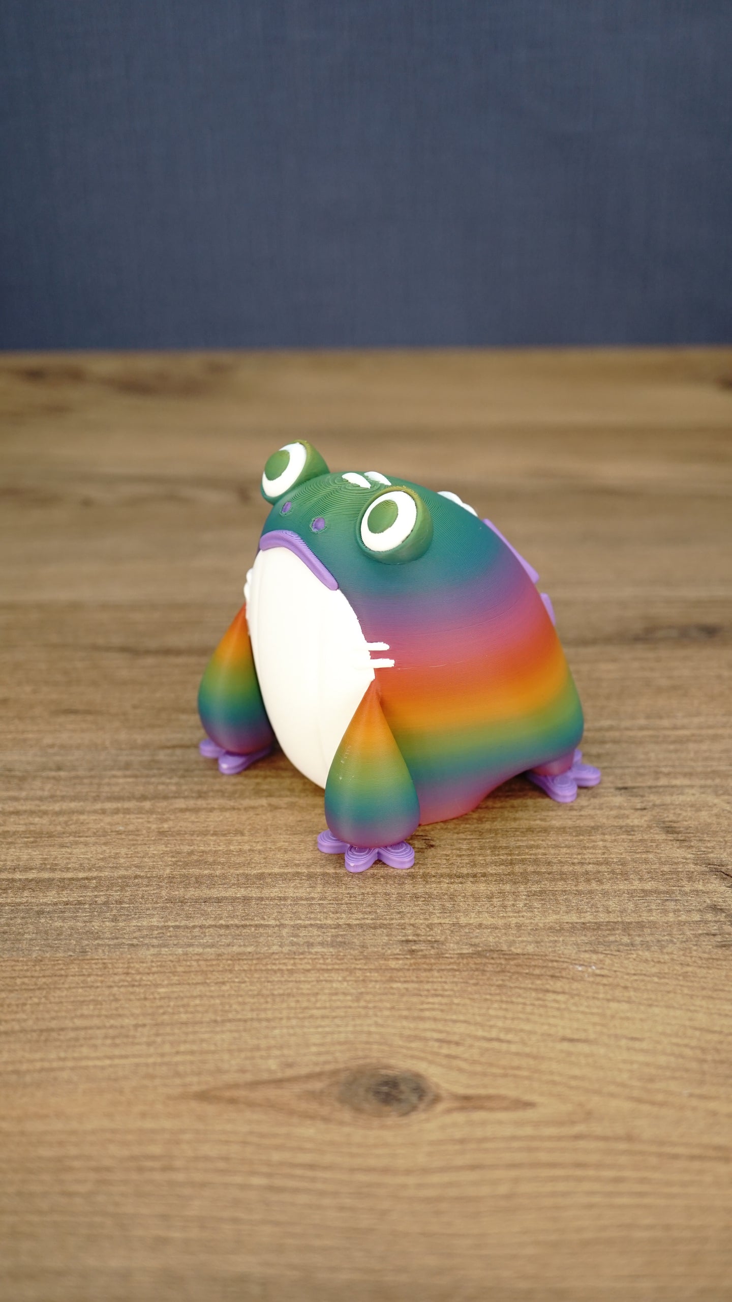 Angled view of a rainbow Frogbert plush figure showing its colorful striped body, white belly, purple feet, and 3D printed texture.