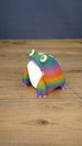 Angled view of a rainbow Frogbert plush figure showing its colorful striped body, white belly, purple feet, and 3D printed texture.