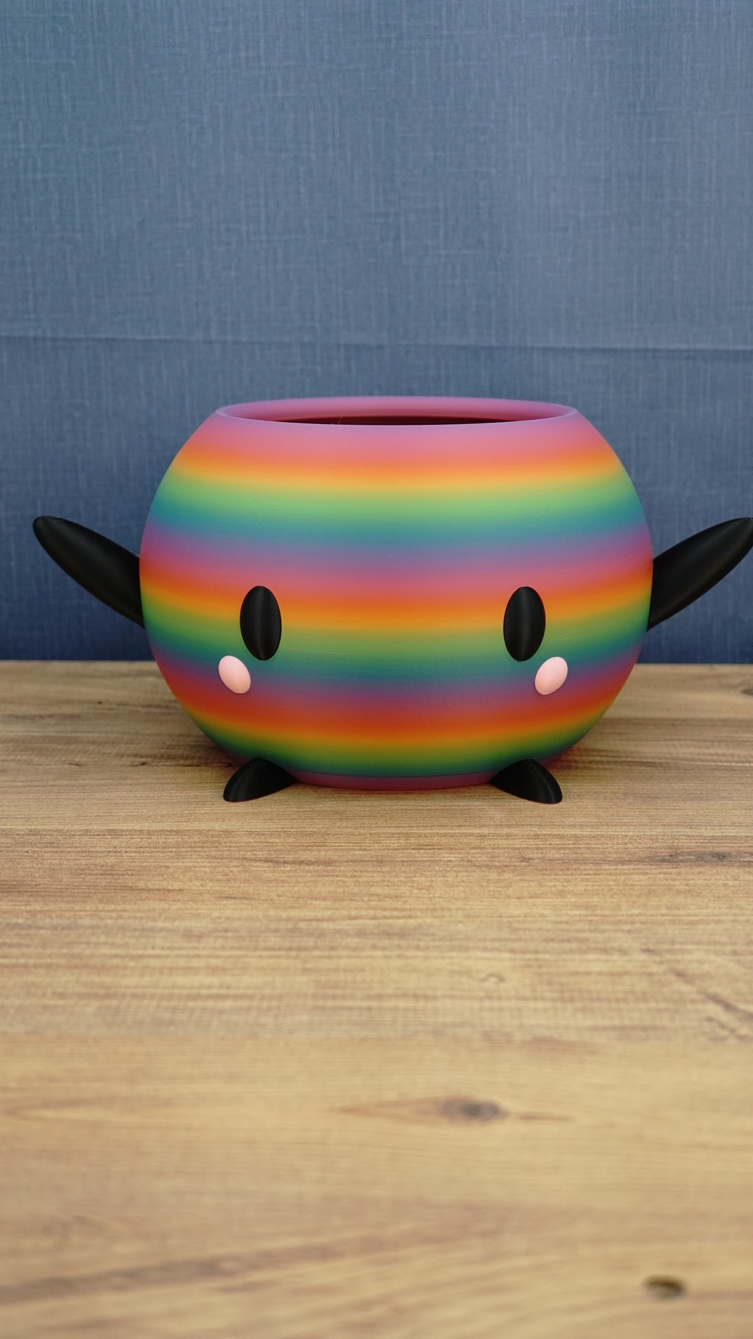 Rainbow-striped Stardew Valley-inspired Junimo planter pot with black arms and feet, oval eyes, and pink cheeks on a wooden tabletop.