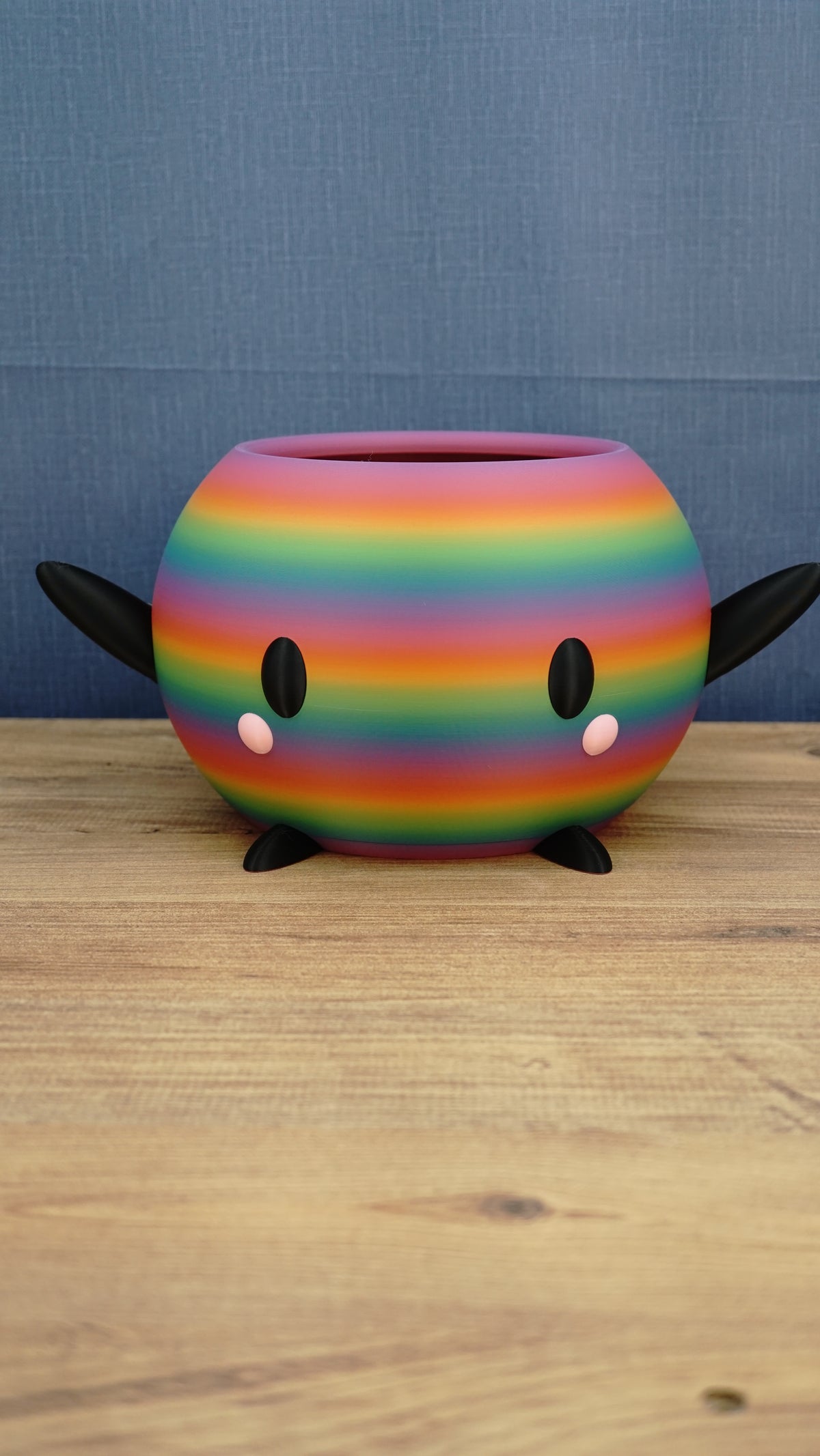 Rainbow-striped Stardew Valley-inspired Junimo planter pot with black arms and feet, oval eyes, and pink cheeks on a wooden tabletop.