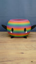 Rainbow-striped Stardew Valley-inspired Junimo planter pot with black arms and feet, oval eyes, and pink cheeks on a wooden tabletop.