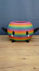Rainbow-striped Stardew Valley-inspired Junimo planter pot with black arms and feet, oval eyes, and pink cheeks on a wooden tabletop.