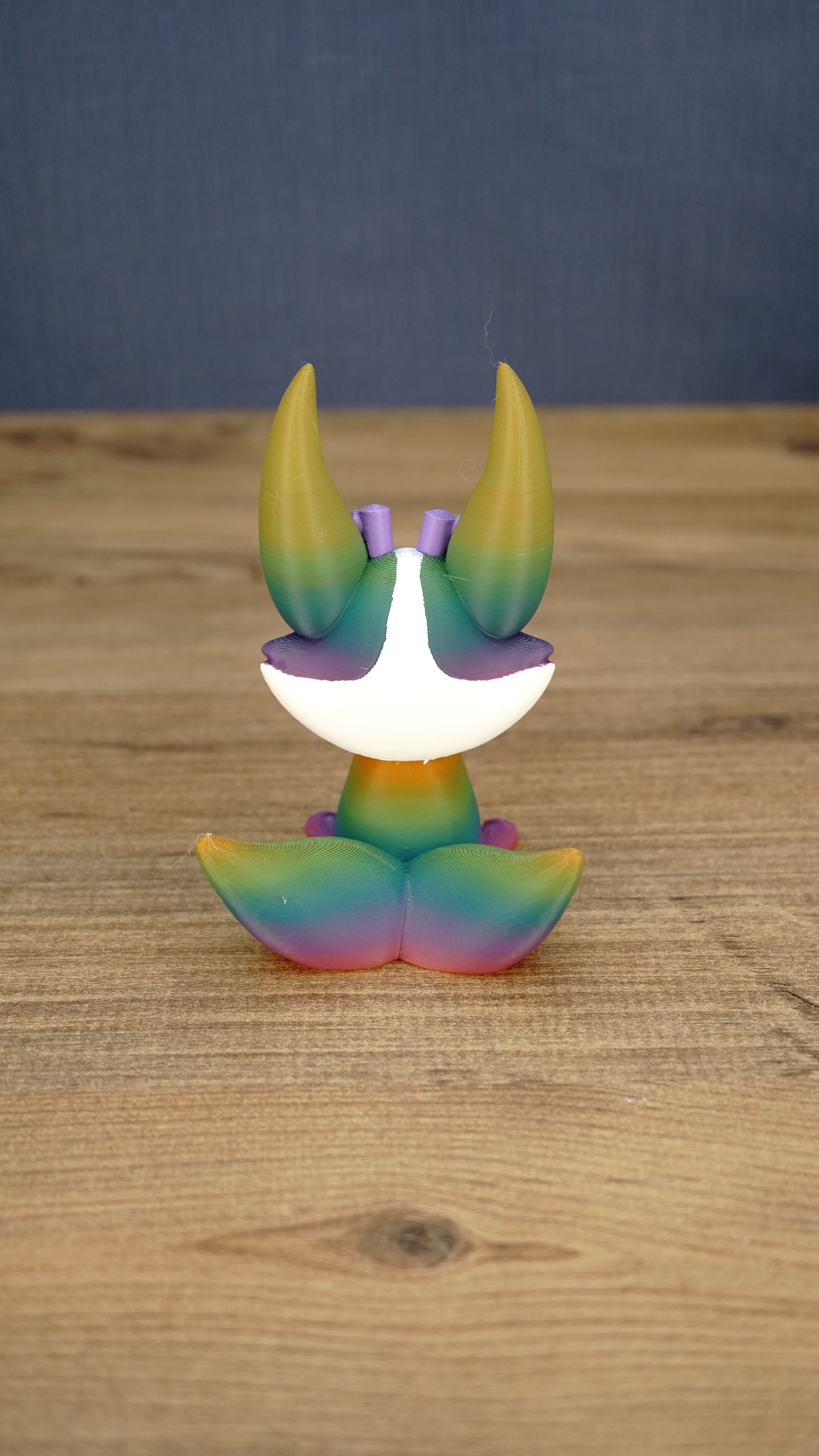 Back view of a rainbow Kitsuu figure showing its large ears, white head marking, and gradient tail colors.