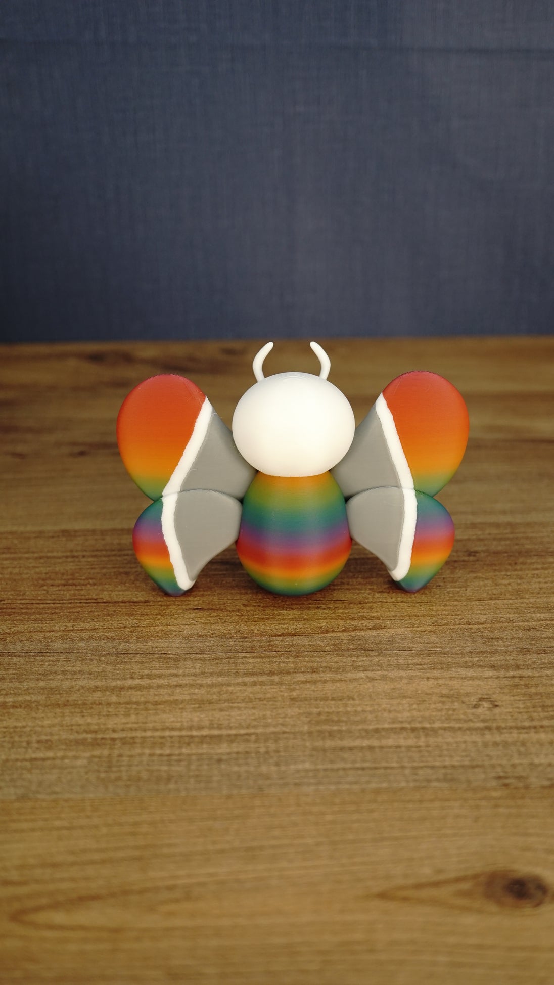 Back view of the Rainbow-Tipped Butterfly Plush figure displaying its symmetrical rainbow wing tips, grey inner wings and rounded multicoloured body.