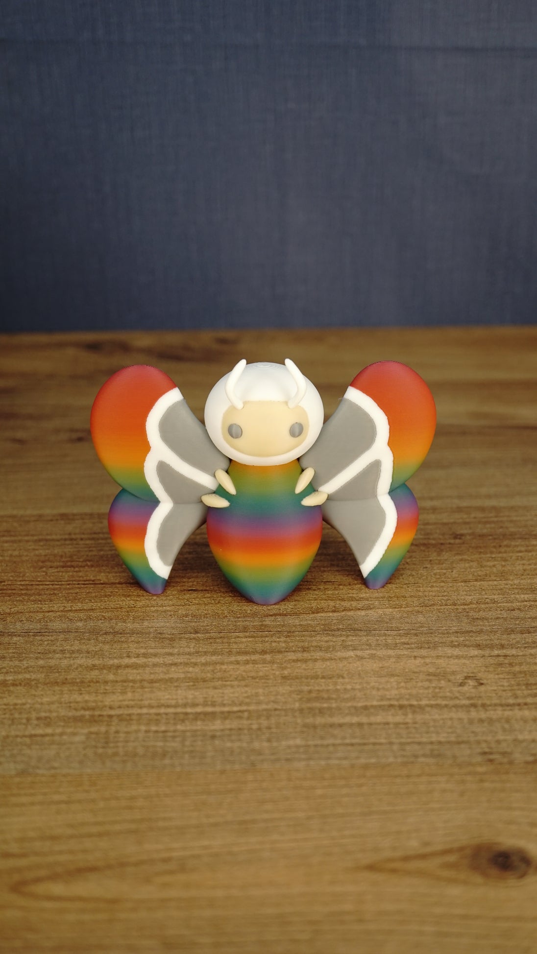 Front view of the Rainbow-Tipped Butterfly Plush figure with a rainbow body, grey and white wings, beige face and curved white antennae