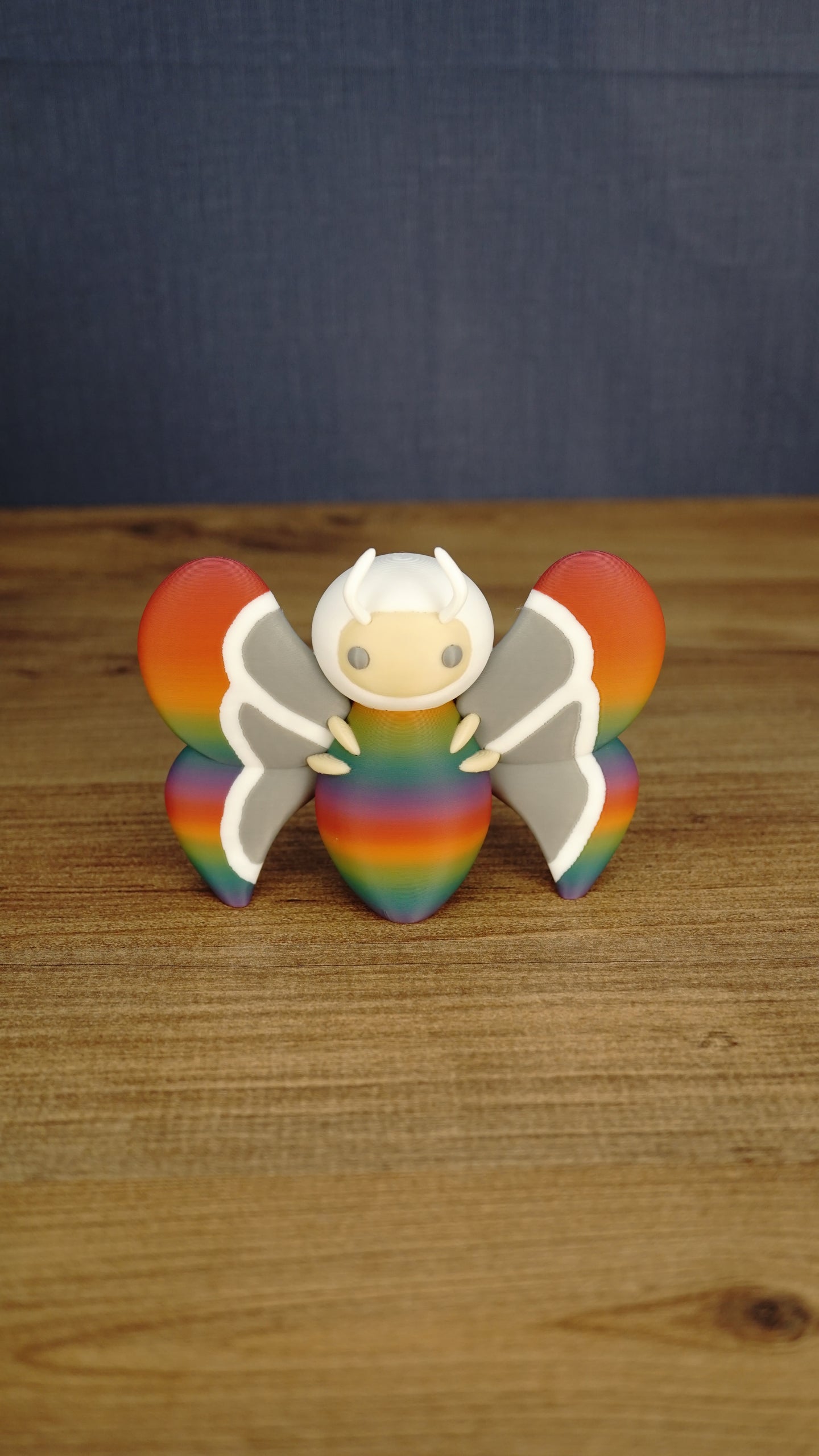 Front view of the Rainbow-Tipped Butterfly Plush figure with a rainbow body, grey and white wings, beige face and curved white antennae