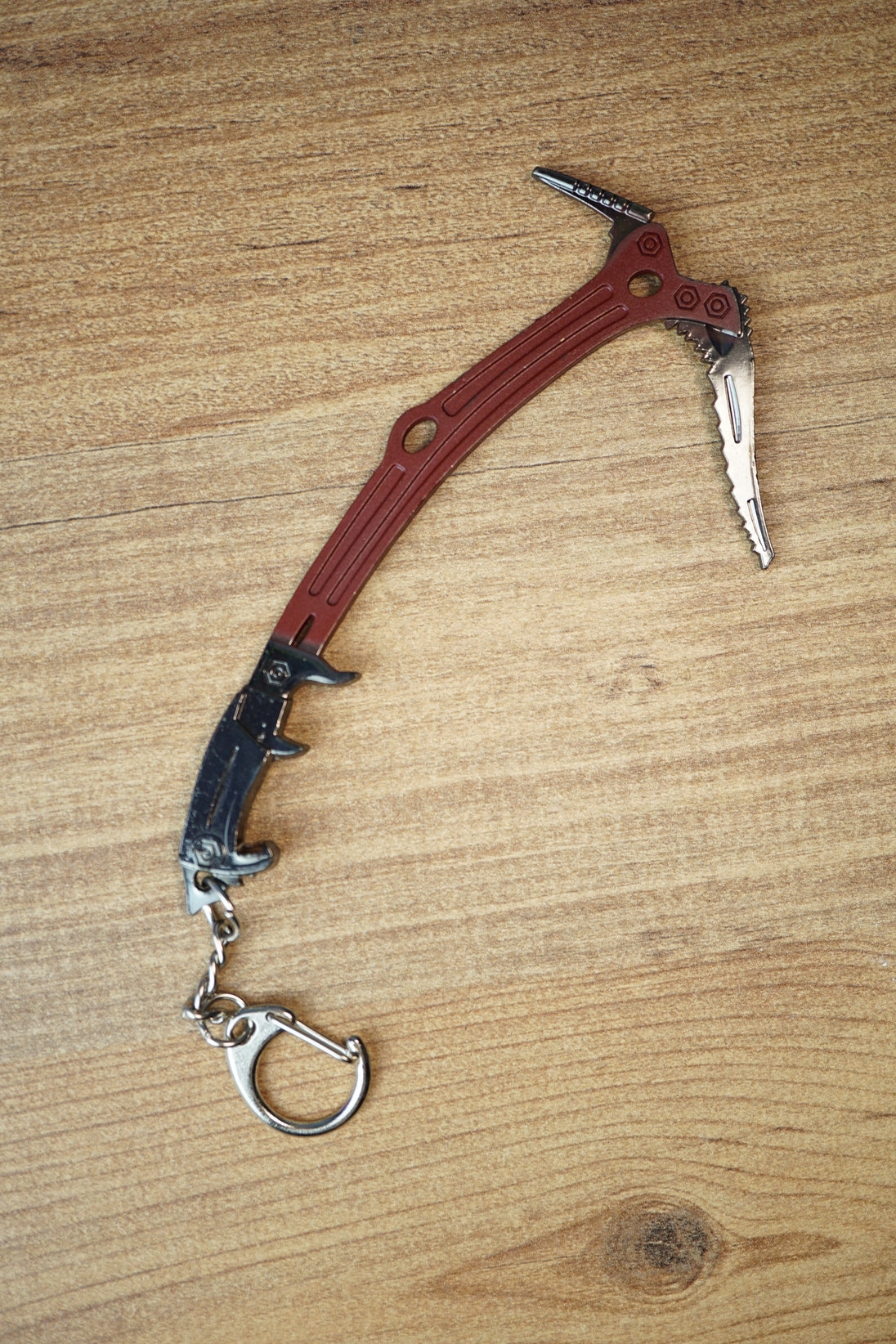 Red Rebel ice pick keyring replica inspired by Tarkov, shown flat on a wooden surface with red handle, black grip, silver pick, and keychain clip.