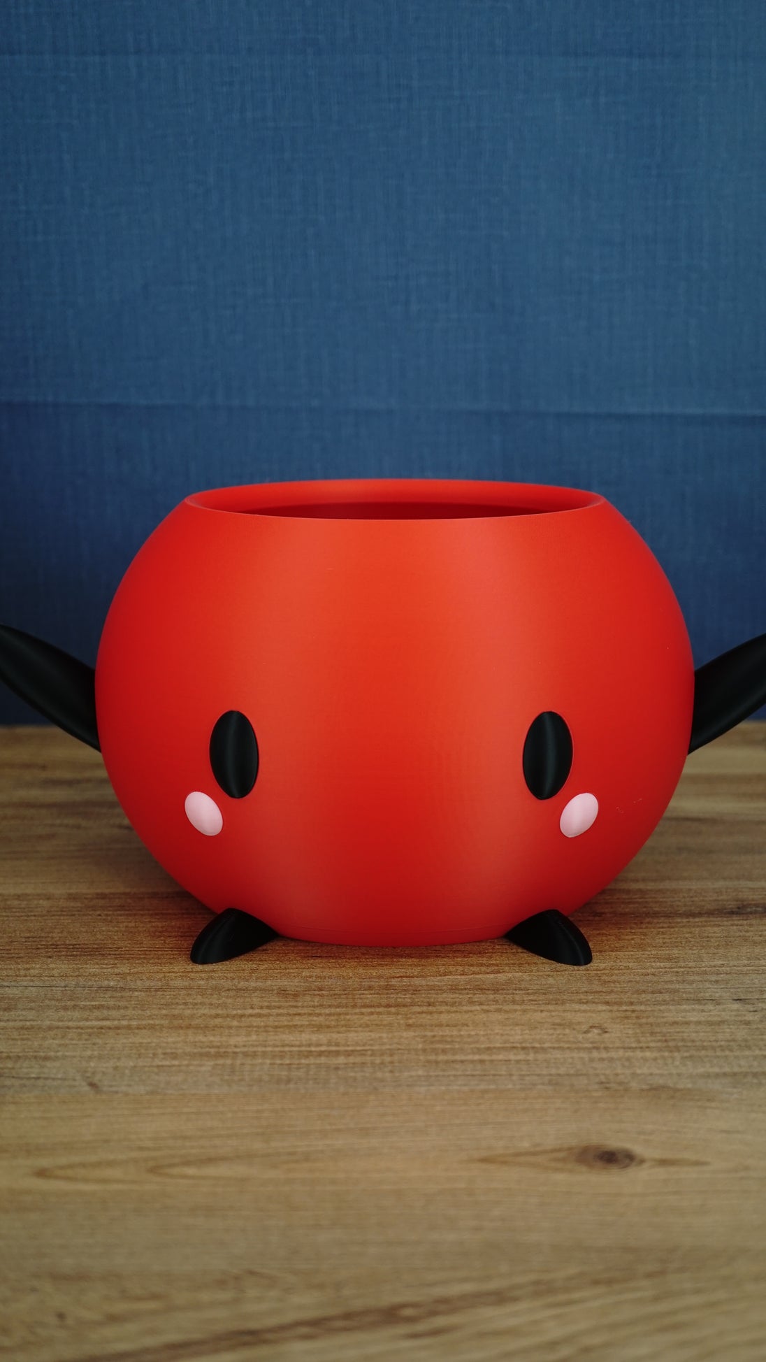 Large red Stardew Valley Junimo planter pot with black arms, black feet, oval black eyes, and pink cheeks on a wooden tabletop.