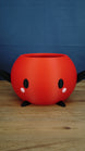 Large red Stardew Valley Junimo planter pot with black arms, black feet, oval black eyes, and pink cheeks on a wooden tabletop.