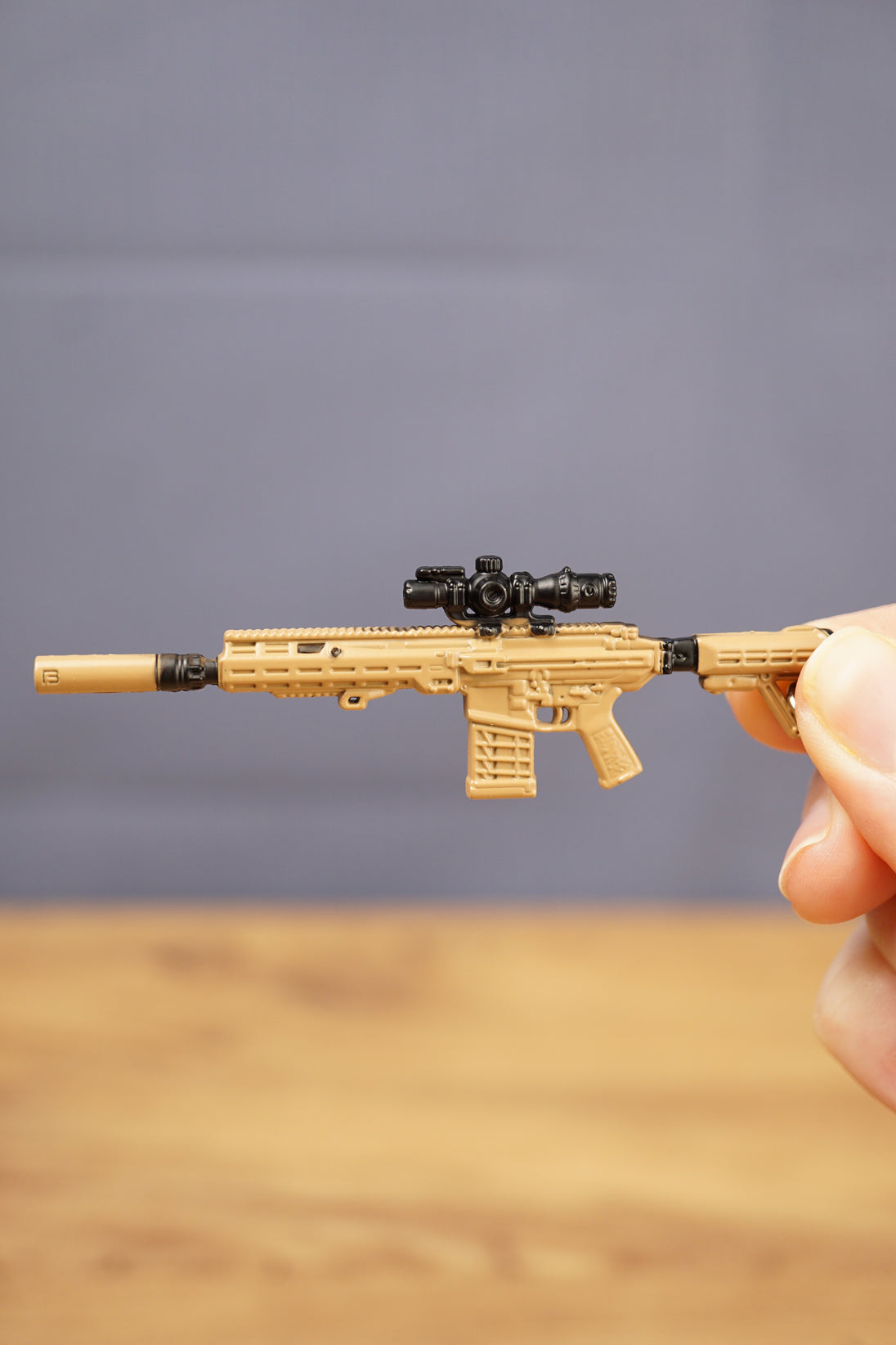 Close-up of tan SIG MCX-SPEAR style miniature rifle keychain with black scope detail, held by hand to show compact size