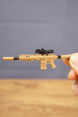 Close-up of tan SIG MCX-SPEAR style miniature rifle keychain with black scope detail, held by hand to show compact size