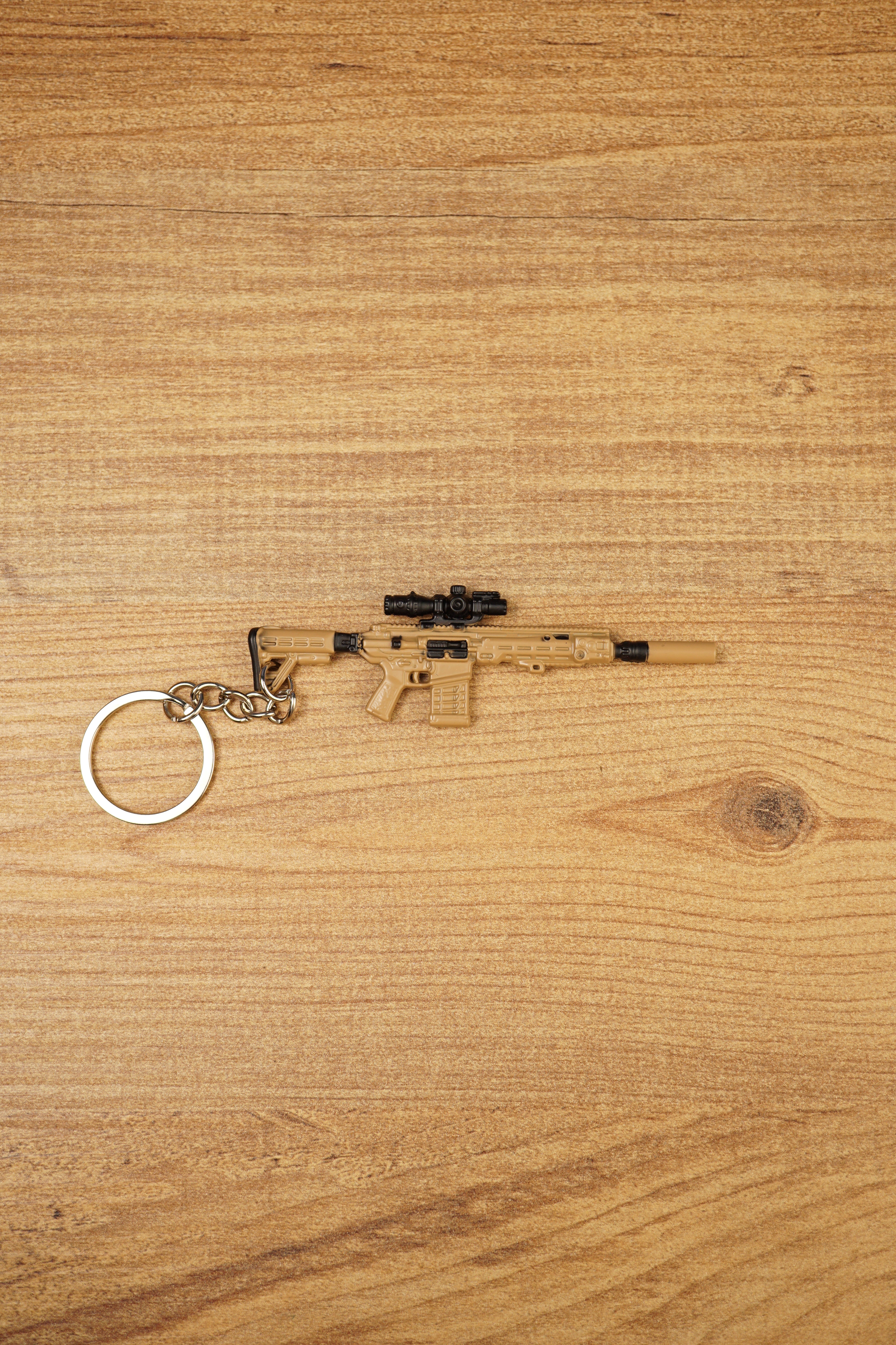 Tan SIG MCX-SPEAR style rifle keychain with black scope and metal keyring laid flat on a wooden surface for tactical FPS fans