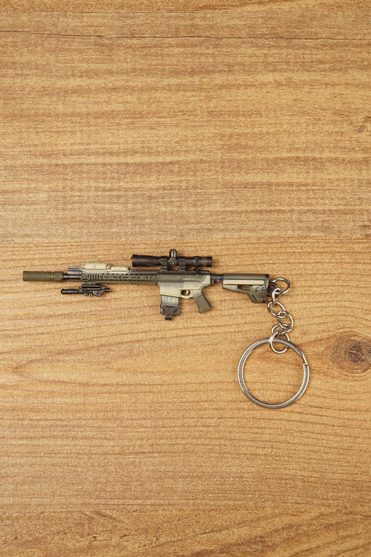 SR-25 DMR rifle keychain laid flat on a wooden surface, showing the tan tactical body, scope, magazine, stock, and metal keyring attachment