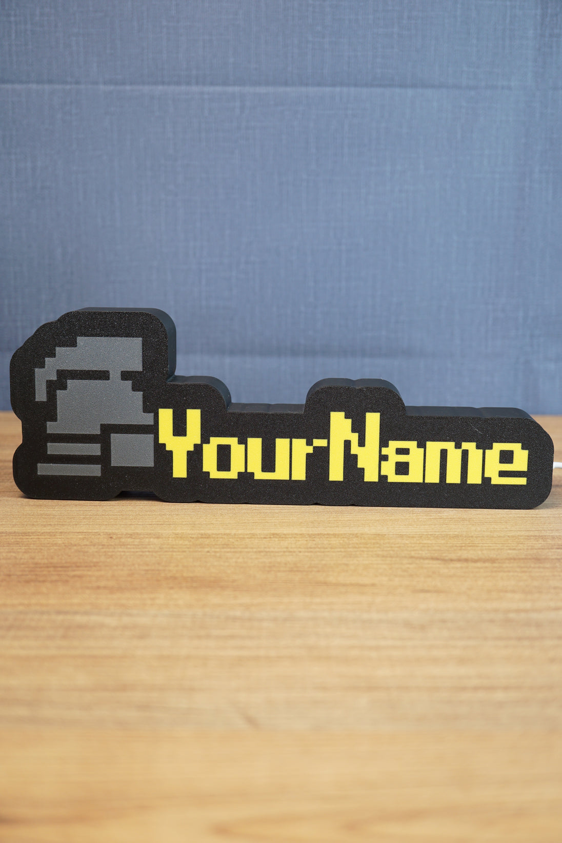 RuneScape-style standard ironman nameplate with a grey helmet icon, yellow custom lettering, and black frame displayed on a wooden desk.