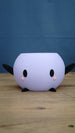 Purple Stardew Valley Junimo planter pot with black arms, feet, eyes, and pink cheeks, shown from the front on a wooden tabletop.
