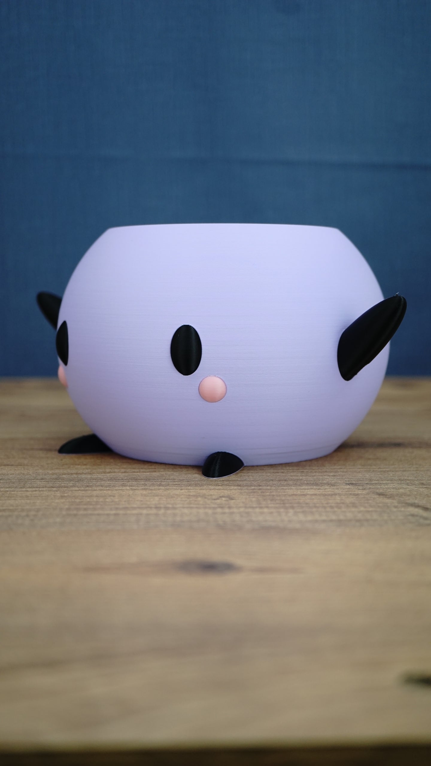 Side view of a purple Stardew Valley Junimo planter pot with black arms, feet, oval eyes, and pink cheeks on a wooden tabletop.