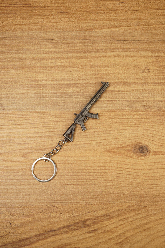 Angled MK47-style metal rifle keychain on wood, ideal as a tactical FPS gamer gift, backpack charm or collectible accessory