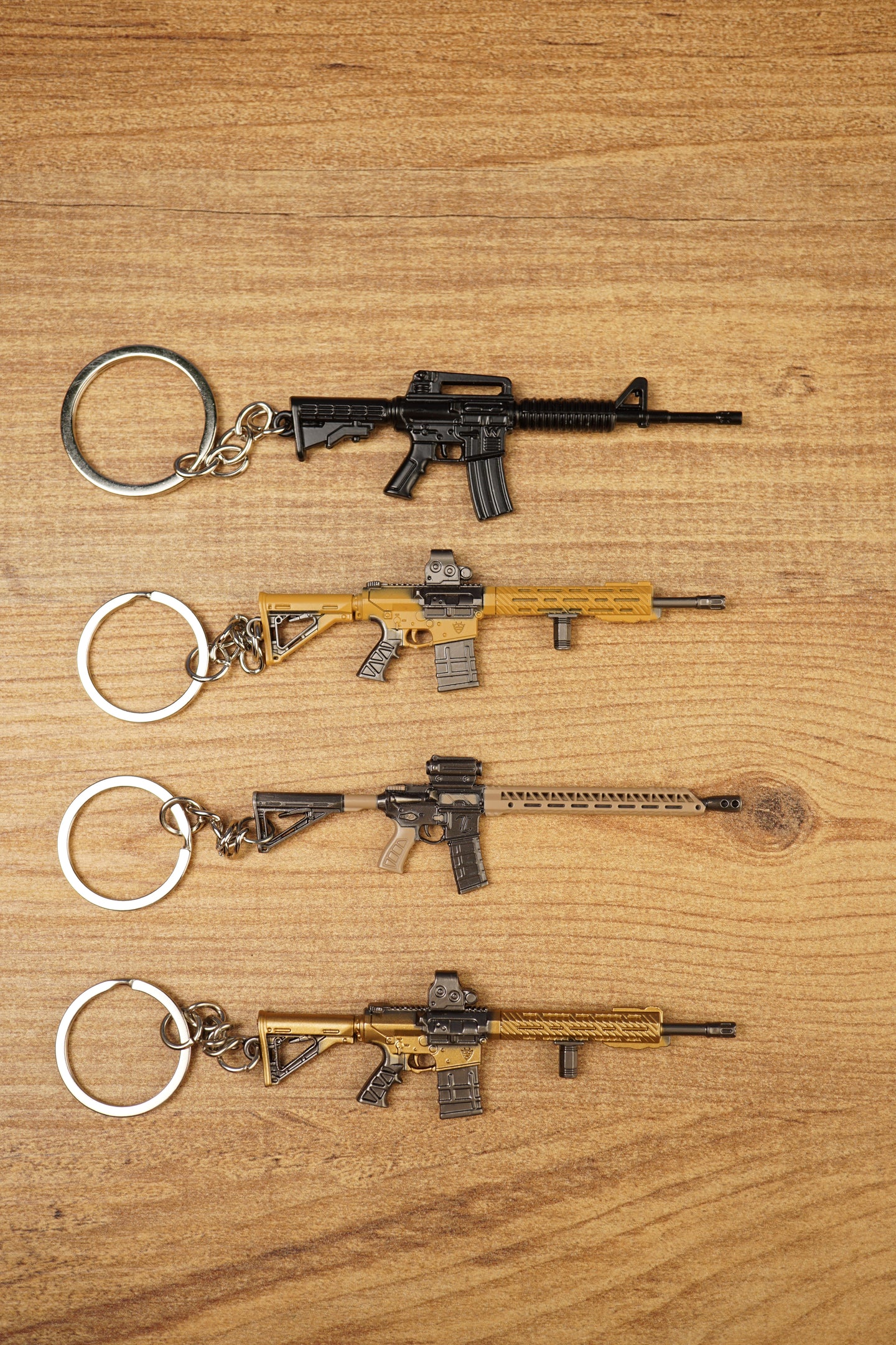 Four miniature tactical rifle keyrings in black, gold and tan styles laid flat on a wooden surface for gamer gift display.