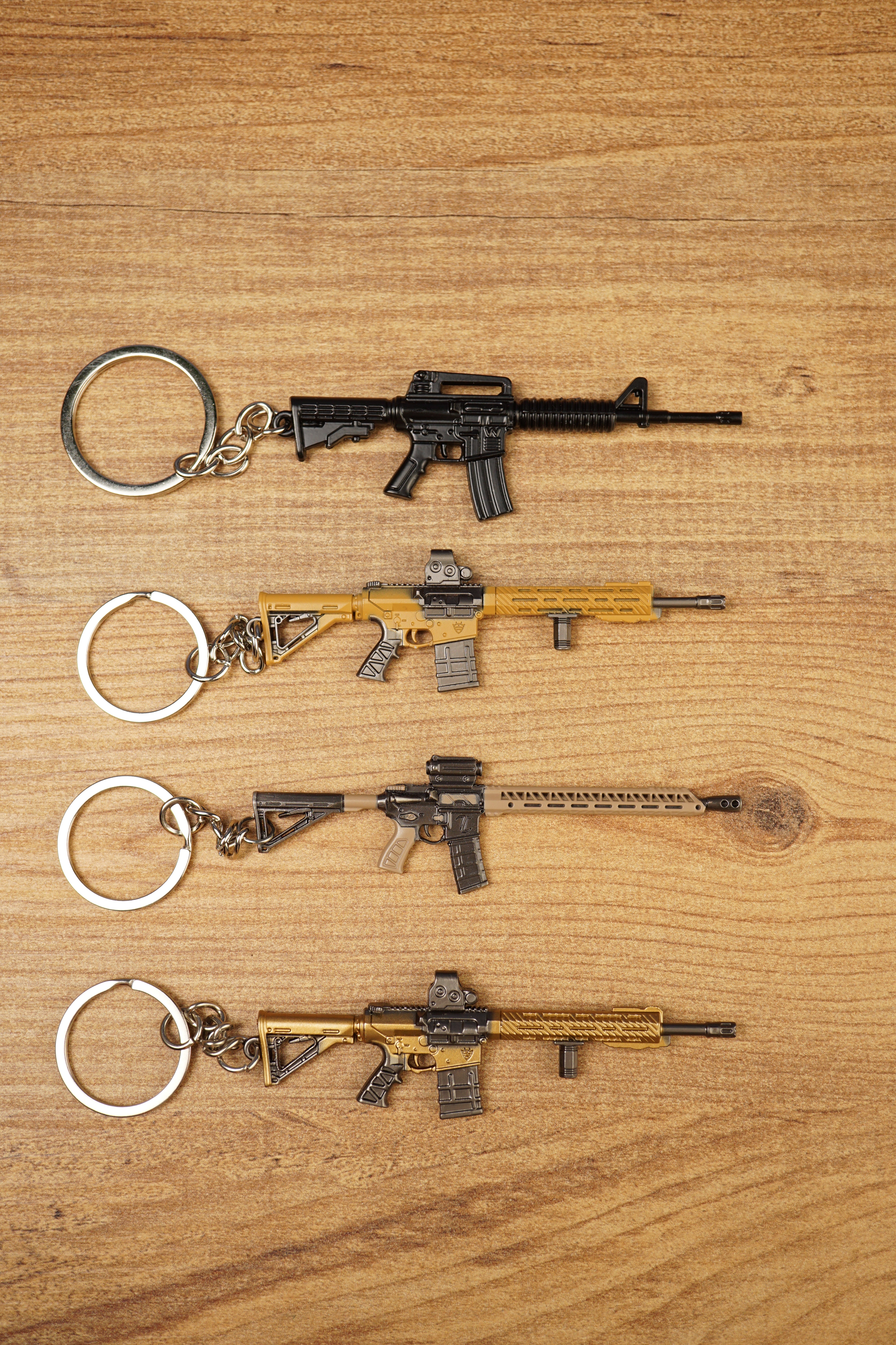 Four miniature tactical rifle keyrings in black, gold and tan styles laid flat on a wooden surface for gamer gift display.