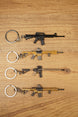 Four miniature tactical rifle keyrings in black, gold and tan styles laid flat on a wooden surface for gamer gift display.