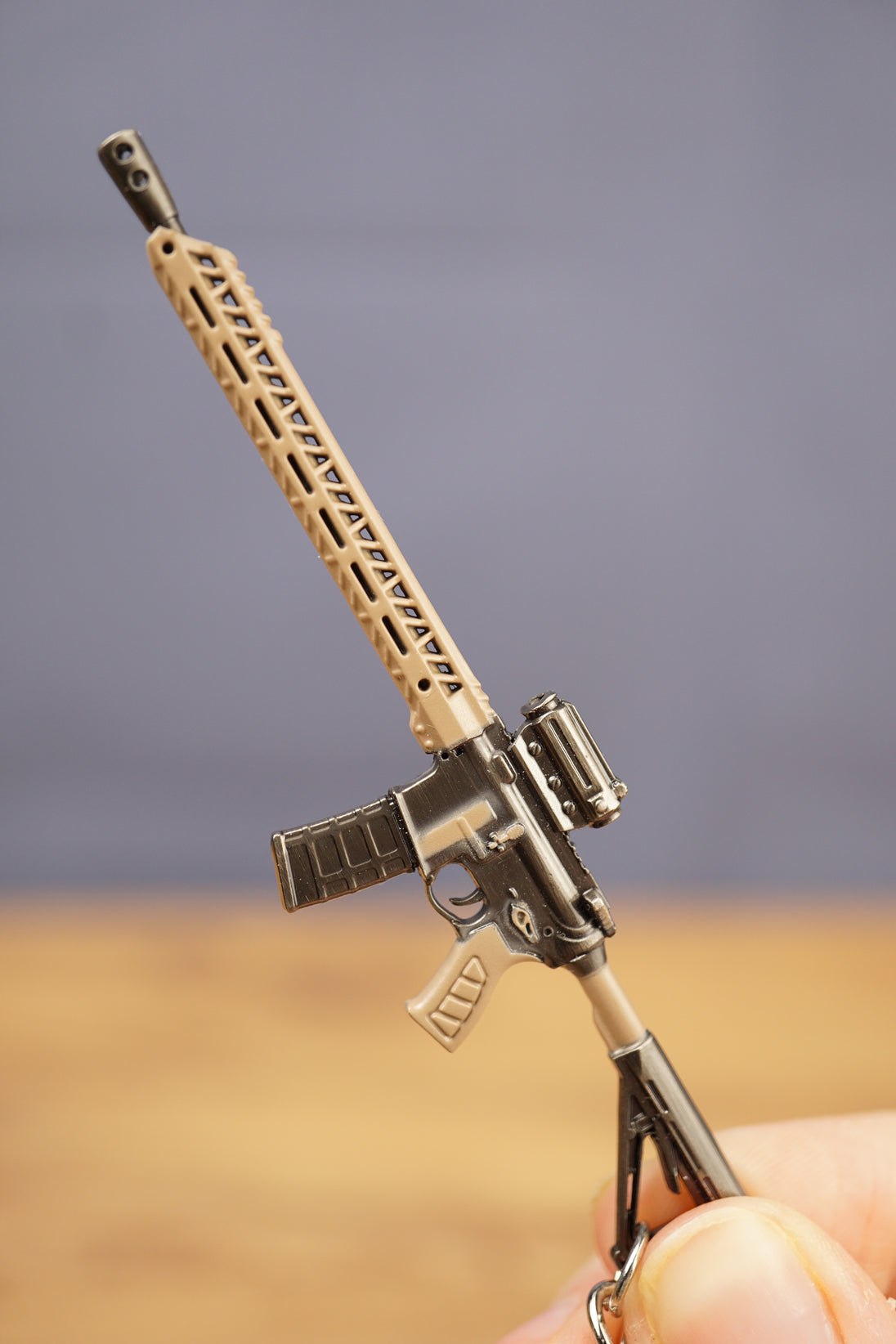 Close-up of tan and black tactical rifle keyring held by fingers, showing long barrel, sight and magazine details.