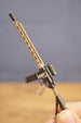 Close-up of tan and black tactical rifle keyring held by fingers, showing long barrel, sight and magazine details.