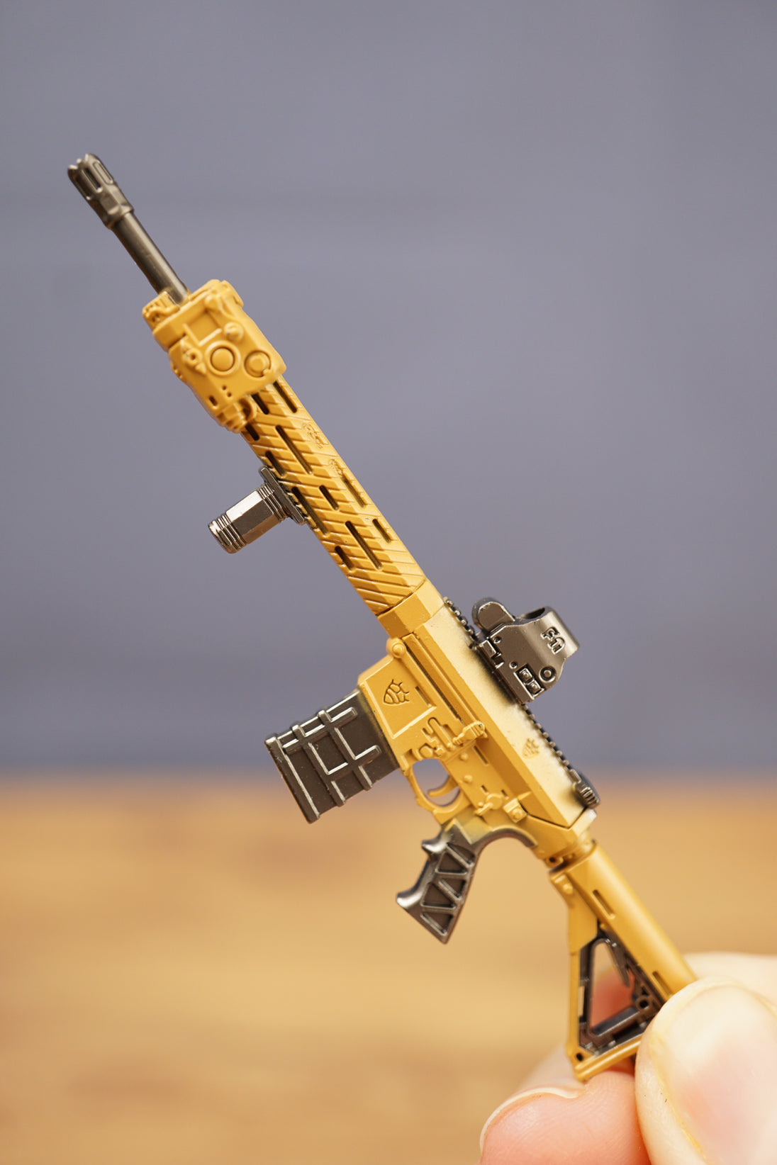 Close-up of tan tactical rifle keychain held between fingers showing detailed barrel, sight, magazine and grip.